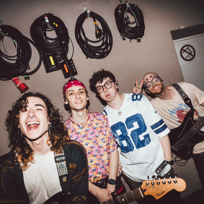 the four of them backstage. nick has long curly brown hair, george is wearing a red bandana and a pink shirt, callum is wearing some sort of sports shirt and his glasses & iain is wearing a graphic tee, their glasses and colourful makeup. when you hover the image, it has a greyscale filter and the album title is visible on it. clicking on this image directs you to their official site!
